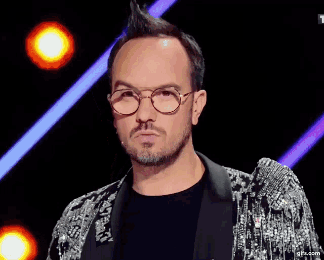 a man wearing glasses and a sequined jacket stands in front of a purple background