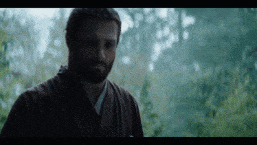 a man with a beard wearing a kimono stands in front of trees