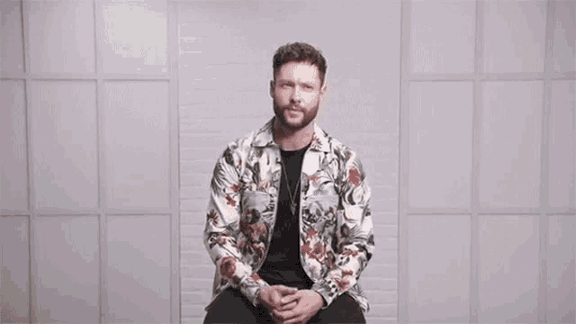 a man in a floral jacket is sitting in front of a wall with the words love conquers all above him