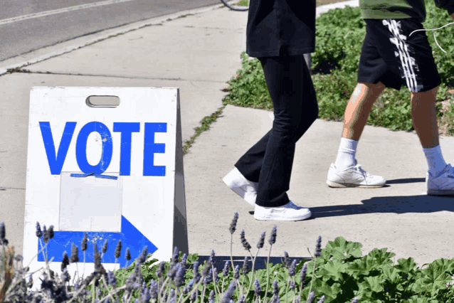 people walk past a sign that says vote on it