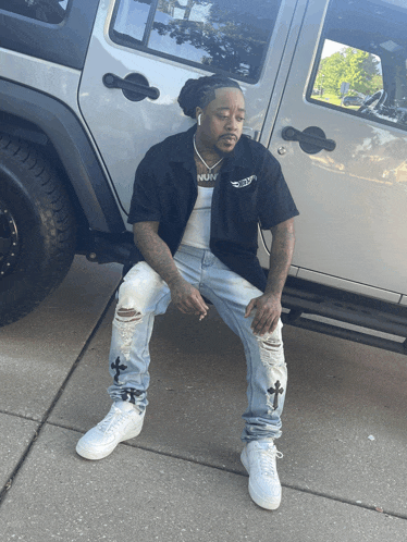 a man wearing a nun shirt sits on the sidewalk in front of a silver jeep