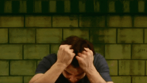 a man holds his head in front of a green brick wall