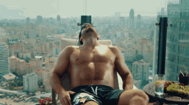 a shirtless man is laying on a balcony with a city in the background