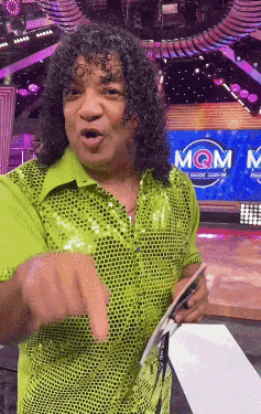 a man in a green sequined shirt is pointing at something in front of a sign that says mqm
