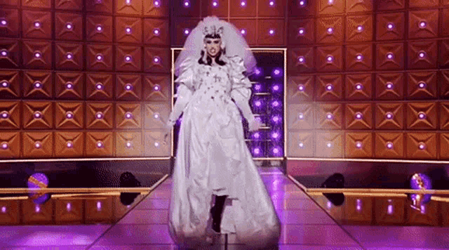 a drag queen in a white bride 's dress is walking down a runway on a stage .