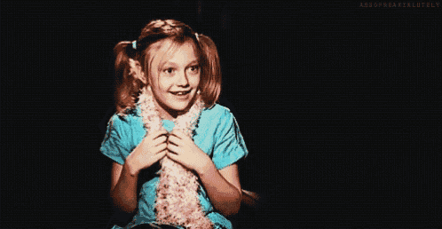 a little girl wearing a blue shirt and a pink scarf smiles while sitting in a chair