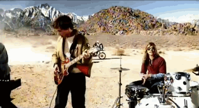 a man is playing a guitar while another man plays drums in front of a mountain .