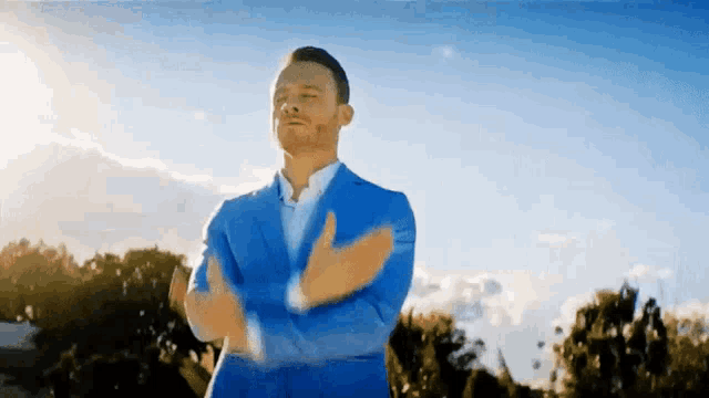 a man in a blue suit stands with his arms crossed in front of a blue sky