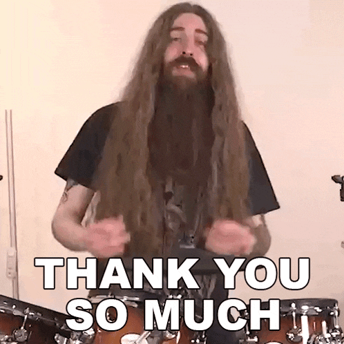a man with long hair and a beard is standing in front of a drum set and saying thank you so much .