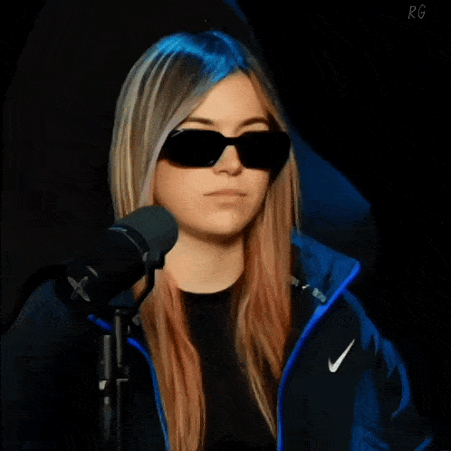 a girl wearing sunglasses and a blue nike jacket stands in front of a microphone