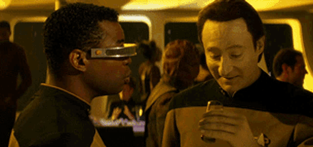a man wearing a pair of futuristic glasses talks to another man holding a glass