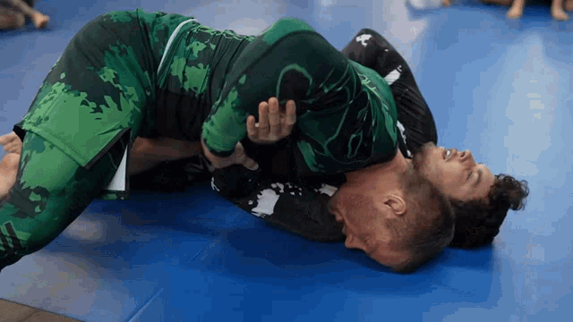 two men are wrestling on a blue mat and one of them is wearing a green uniform