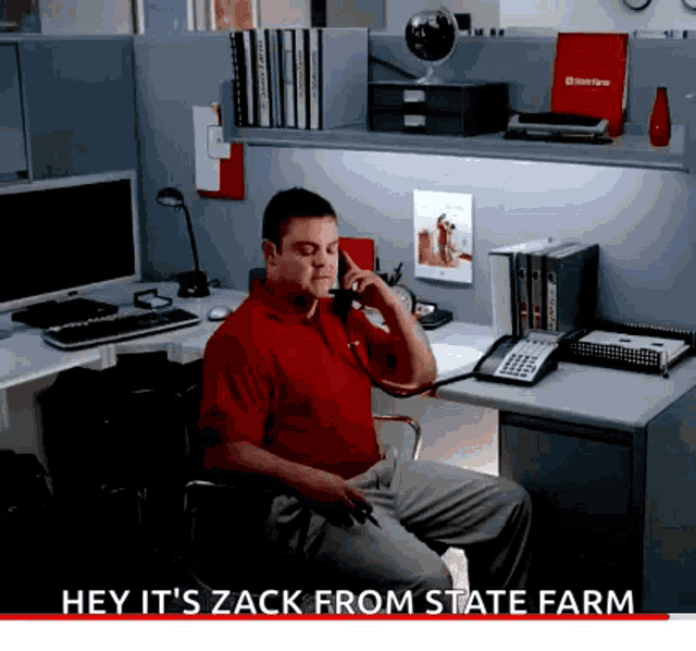 a man in a red shirt sits at a desk talking on the phone