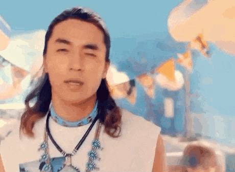 a man with long hair and a necklace is standing in front of a beach .