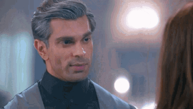 a man in a suit and black turtleneck is looking at a woman in a room .