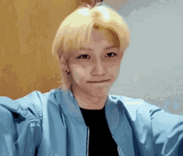 a young man with blonde hair and a blue jacket is taking a selfie .