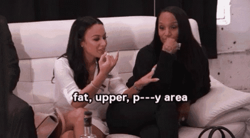 two women are sitting on a couch talking to each other . one of the women is holding a bottle of alcohol .
