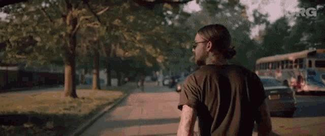 a man with long hair and sunglasses is walking down a street with a bus in the background .