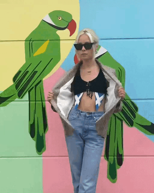 a woman is standing in front of a painting of green parrots and holding her jacket up