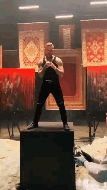 a man in a black tank top is standing on a box in front of a wall of paintings .