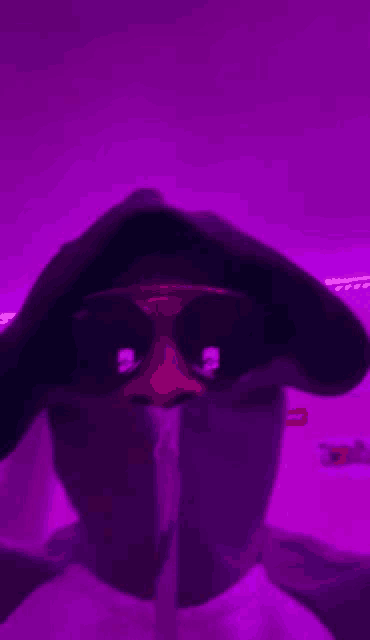a person wearing sunglasses and a hoodie is taking a selfie in front of a purple background .