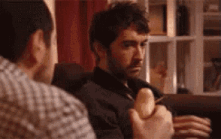 a man is sitting on a couch eating a banana while another man watches .