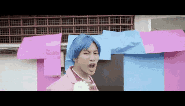a man with blue hair and a pink jacket is standing in front of a wall covered in pink and blue papers .