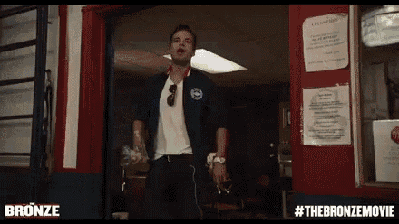 a man in a blue jacket is standing in front of a red door with #thebronzemovie written on it