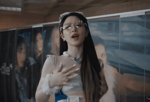 a woman wearing glasses and a pearl necklace stands in front of a wall with a picture of a woman behind her