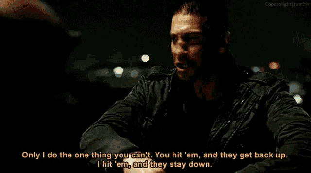 a man in a black jacket is holding a gun and talking to another man in a dark room .
