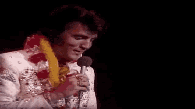 elvis presley is singing into a microphone on a stage while wearing a lei around his neck .
