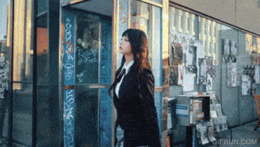 a woman in a suit stands in front of a building with graffiti on it and a gifrun.com logo in the corner