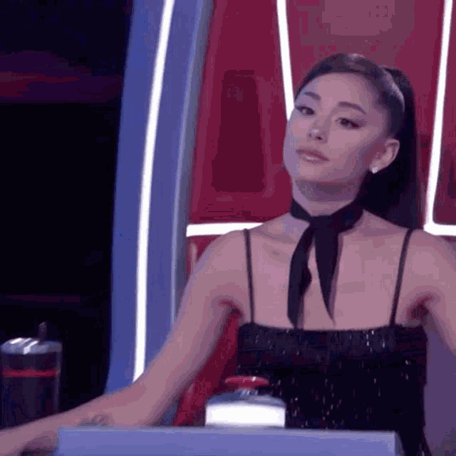 ariana grande is wearing a black dress and a black tie while sitting at a desk .