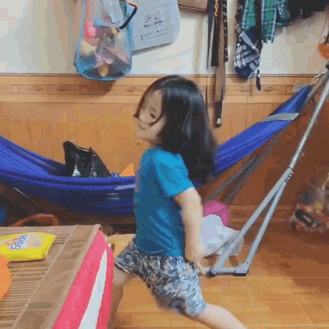 a little girl in a blue shirt and shorts is dancing in front of a hammock .