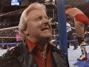 a man in a red shirt and black jacket is standing in a wrestling ring with a wrestler in the background .