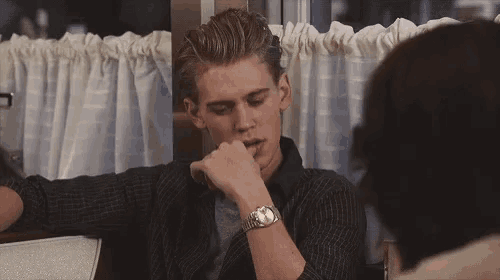 a man wearing a watch is sitting at a table in a diner talking to another man .