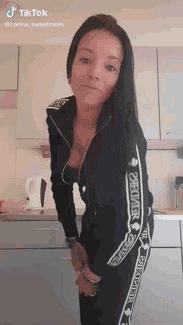 a woman is standing in a kitchen wearing a black tracksuit that says delivery on the sleeves