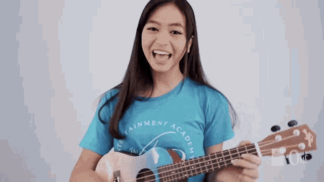 a girl wearing a blue shirt that says entertainment academy is playing a guitar