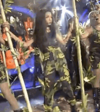 a woman in a camouflage outfit is standing in front of a group of people holding bamboo poles .