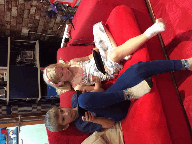 a boy and a girl laying on a red couch with a tablet