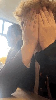 a man in a suit is covering his face with his hands while sitting at a table .