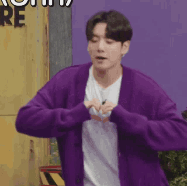 a man wearing a purple cardigan and a white shirt is making a heart with his hands .
