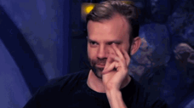 a man with a beard is smiling while sitting in a dark room with his hand on his chin .