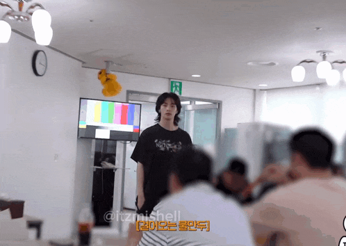 a man in a black t-shirt stands in front of a television with a green exit sign on the wall