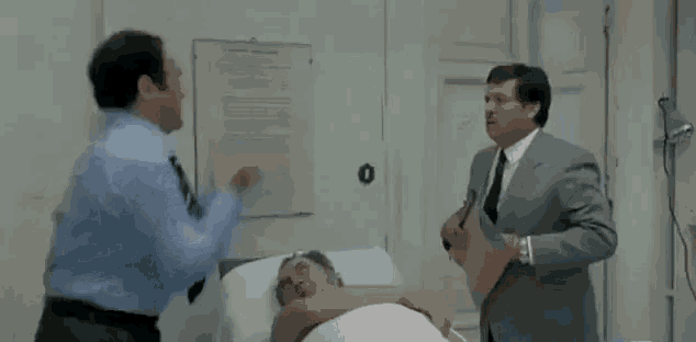 two men are standing around a man in a hospital bed .