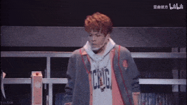 a young man wearing a hoodie and a headband is standing on a stage in front of a bookshelf .