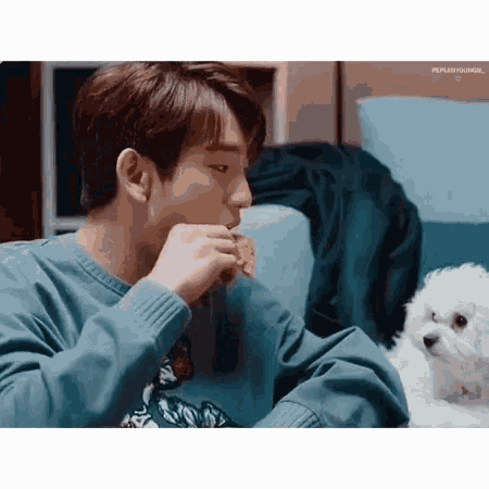 a young man is sitting on a couch eating a piece of food while a small white dog looks on .