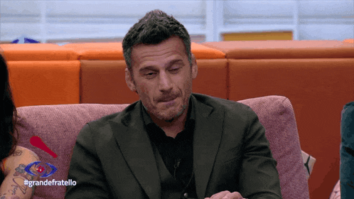 a man in a suit sits on a couch with the hashtag #grandefratello