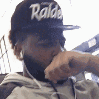 a man wearing a raiders hat is covering his mouth with his hand while listening to music .