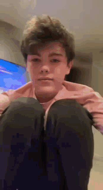 a young boy in a pink shirt and black pants is squatting down in front of a tv .